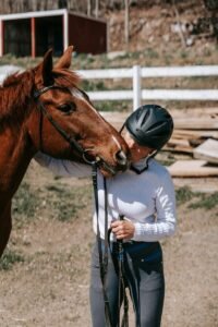 A woman wearing a riding helmet affectionately kisses a horse outdoors on a sunny day.
